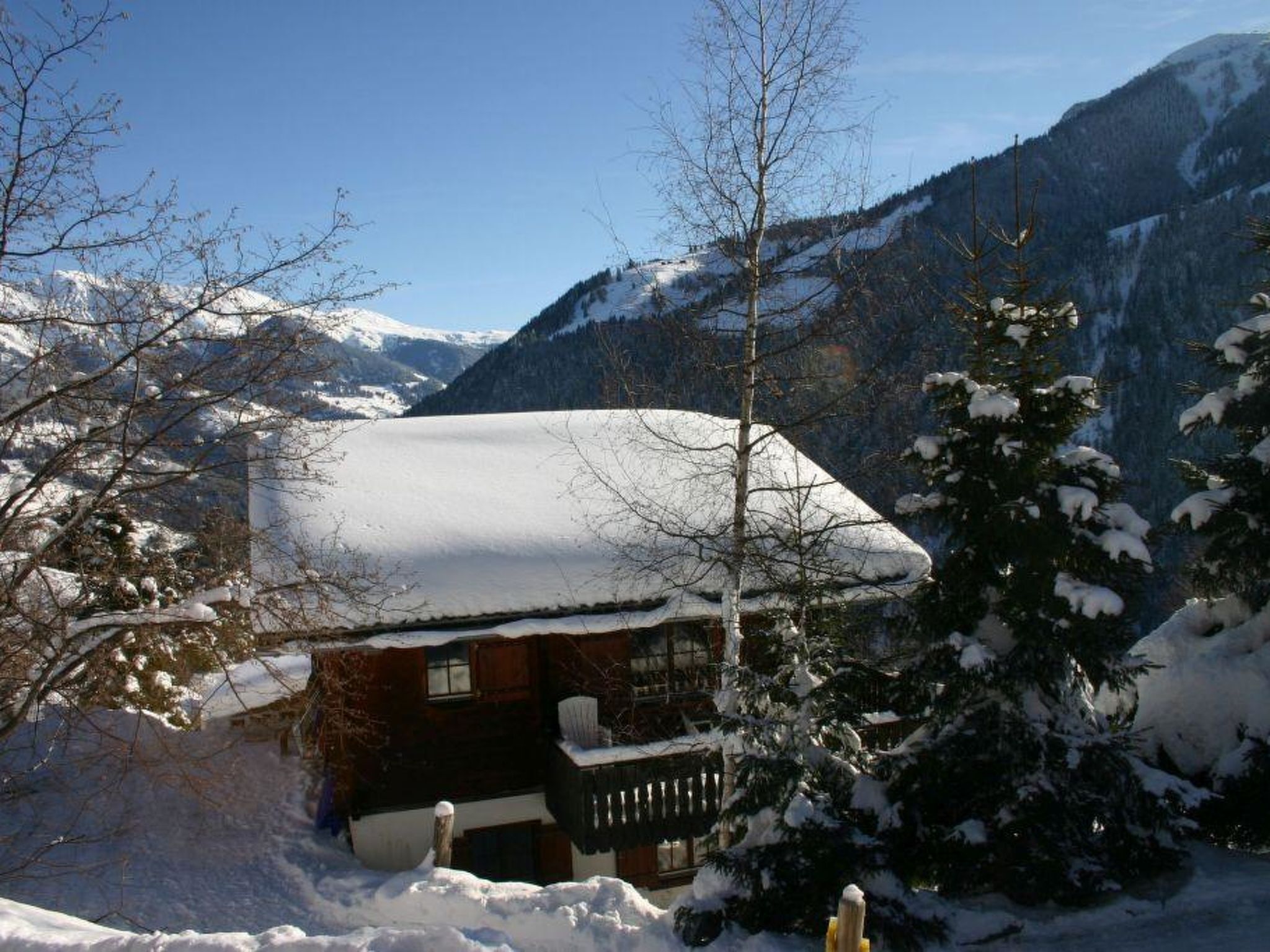 Una cabaña con techo nevado, rodeada de árboles y montañas cubiertas de nieve.
