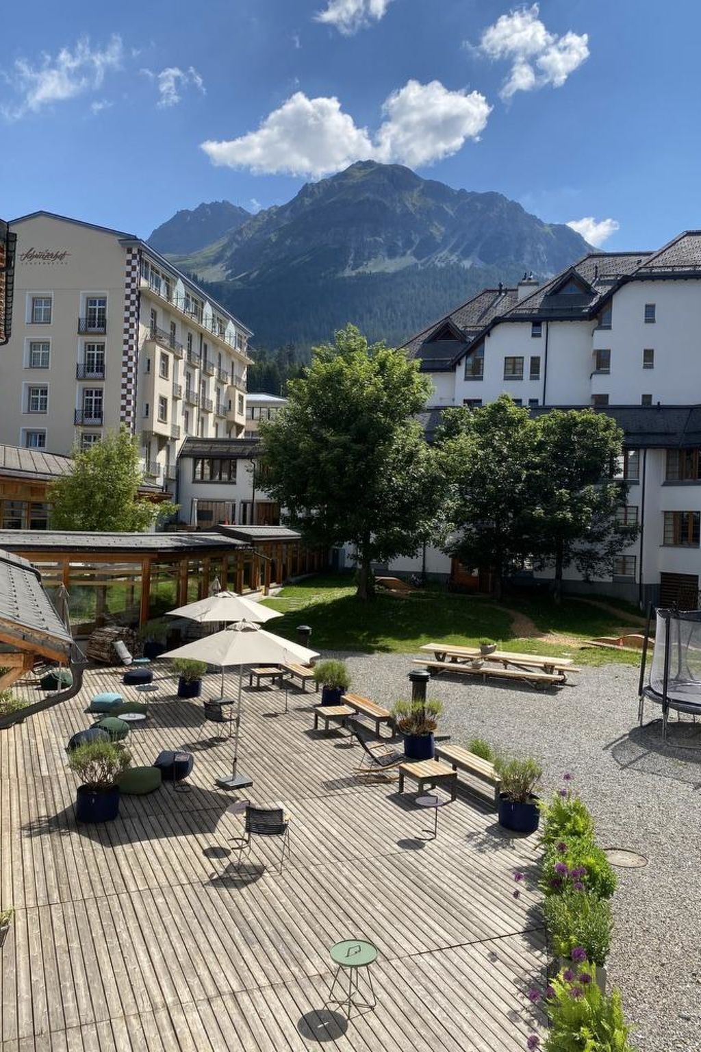 Eine Holzterrasse mit Sitzgelegenheiten im Freien, umgeben von Gebäuden und einem Berg im Hintergrund.