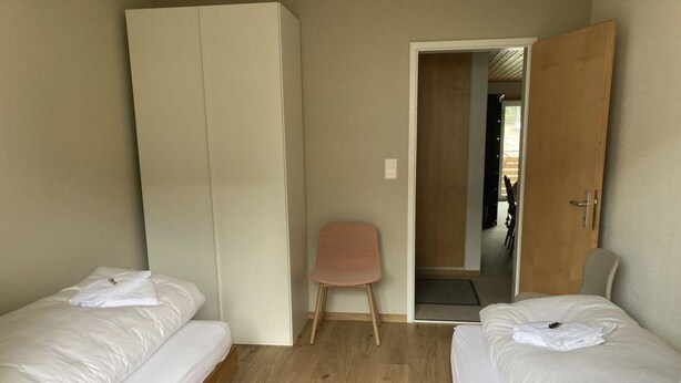Una habitación con dos camas individuales, un armario de madera, una silla pequeña y una puerta que da a otra habitación.
