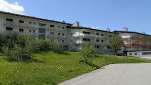 Un edificio residencial de varios pisos con balcones, un área verde con flores amarillas y un cielo despejado.
