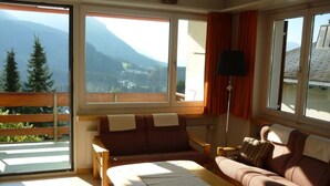 A room with a sofa, a table, and a view of mountains through large windows. - Haus Curver Bächtold Appartement/Fewo,   5-Bettwohnung (4.5 Zimmerwohnung) by Interhome (Valbella)