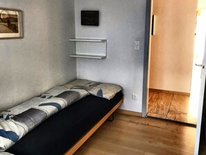 A small, single-bed room with a wooden floor, a wall-mounted shelf, and a door leading to another room. - Haus Curver Bächtold Appartement/Fewo,   5-Bettwohnung (4.5 Zimmerwohnung) by Interhome (Valbella)