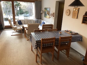 Ein gemütliches Wohnzimmer mit einer Couch, Sesseln, einem Esstisch mit kariertem Tischläufer und Blick ins Freie.