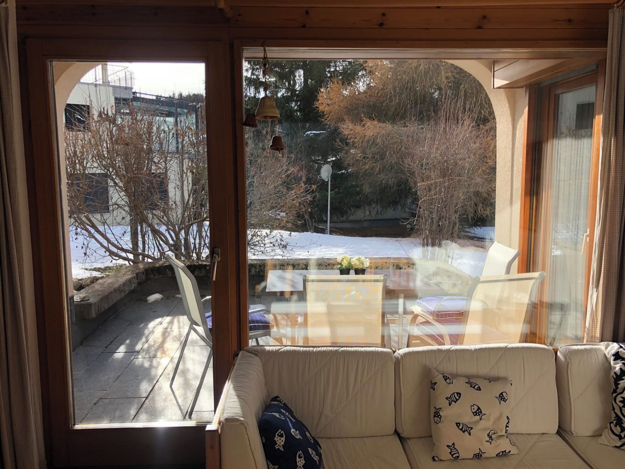 Une pièce comprenant un canapé, une table et donnant sur un espace extérieur enneigé, avec des arbres et un bâtiment en arrière-plan.