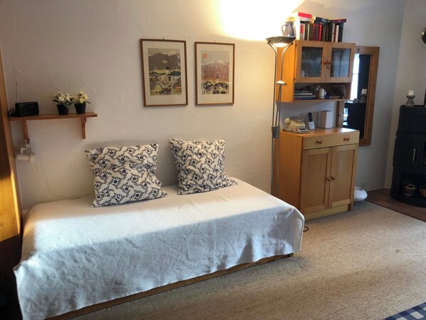 Un dormitorio con una cama, dos almohadas, un armario de madera, una estantería con libros y dos cuadros en la pared.