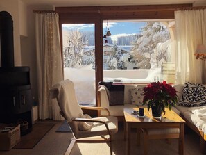 Ein gemütliches Wohnzimmer mit einer Couch, einem Holztisch und einem Sessel, einem Kamin und Blick auf eine verschneite Landschaft.