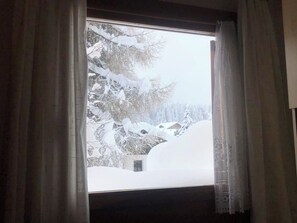 A window with curtains partially drawn, revealing a snowy landscape with trees and buildings outside. - Auf den Böden Haus B Appartement/Fewo,   Auf den Böden Haus B by Interhome (Parpan)
