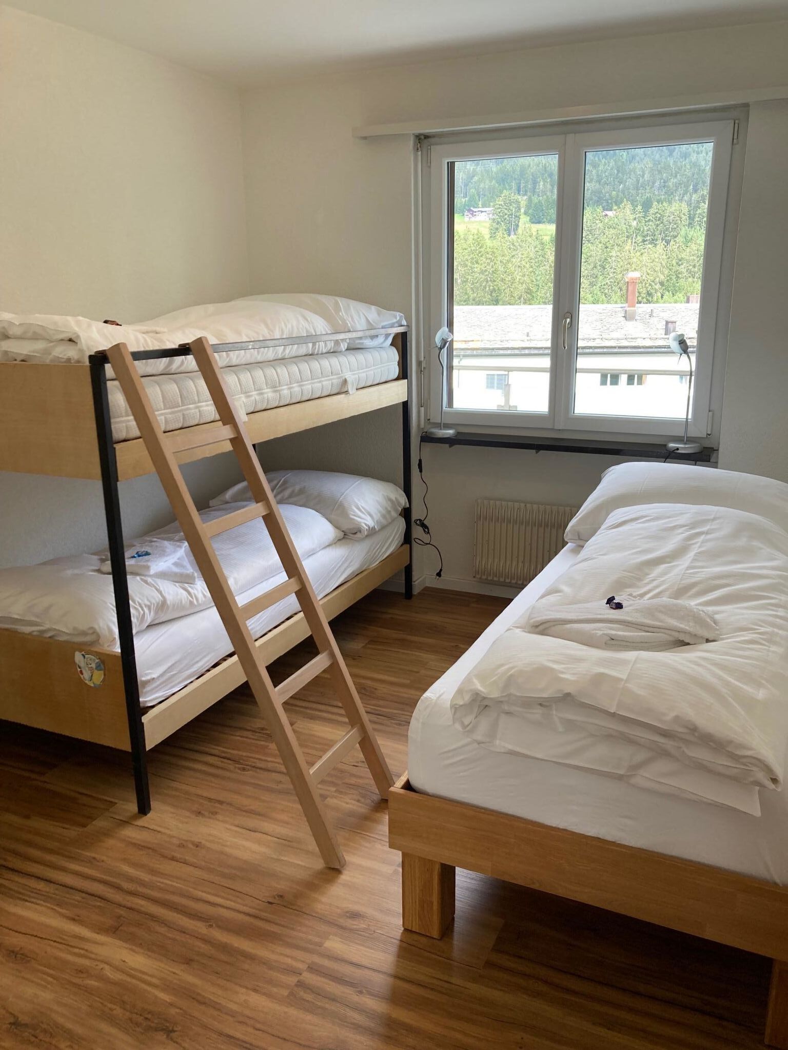 A room with a bunk bed, a single bed, a wooden ladder, and a window with a view of greenery.