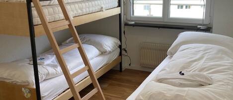 A room with a bunk bed, a single bed, a wooden ladder, and a window with a view of greenery.