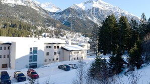 A snow-covered parking lot with several cars, a building with multiple windows and a flat roof, and a mountainous backdrop. - Haus Montana 3/4 Appartement/Fewo,   7-Bettwohnung by Interhome (Lenzerheide)