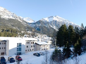 Ein verschneiter Parkplatz mit mehreren Autos, ein Gebäude mit vielen Fenstern und einem flachen Dach sowie eine gebirgige Kulisse.