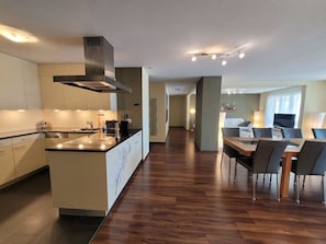 Eine moderne Küche mit Kochinsel, ein Essbereich mit Tisch und Stühlen sowie ein Wohnzimmer mit Sofa und Fernseher.