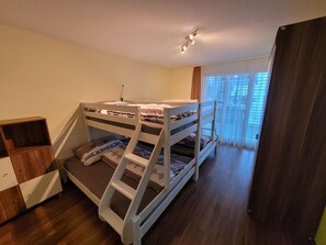 Ein Schlafraum mit Etagenbetten, Holzboden, einem kleinen Holzschrank und einem Fenster mit Vorhängen.