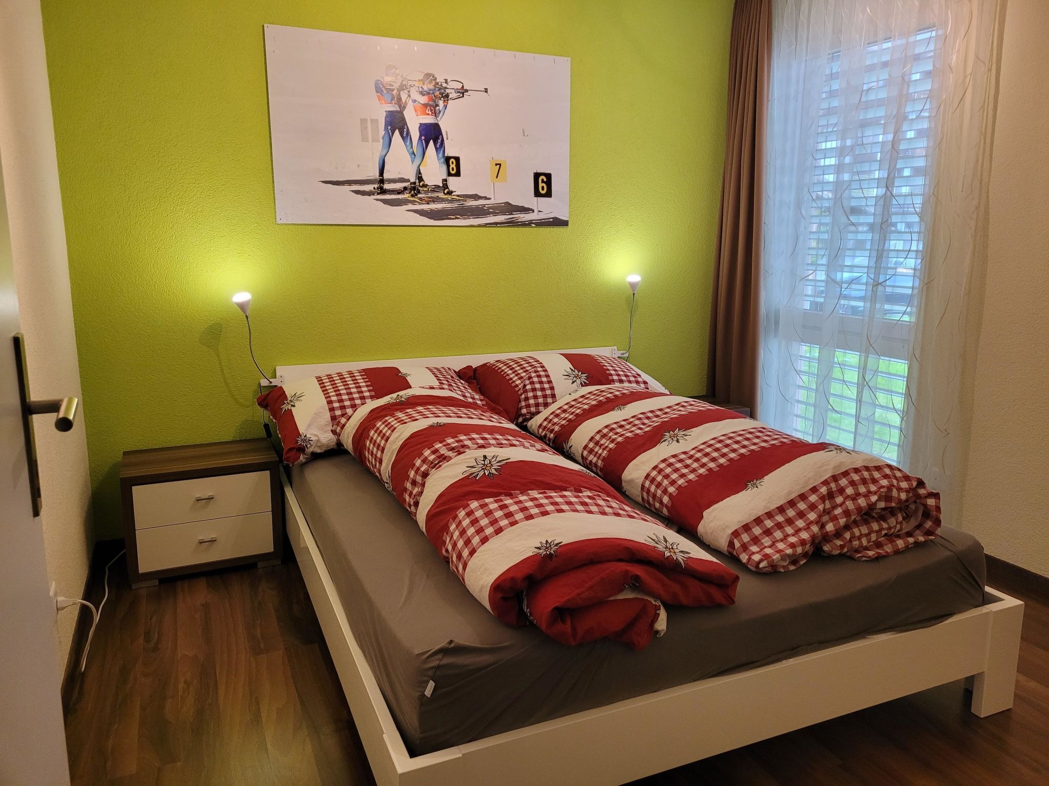 Ein Einzelbett mit rot-weißer Karodecke, ein Nachttisch mit Lampe sowie ein an der Wand hängendes Bild von einer Person in Skiklamotten.