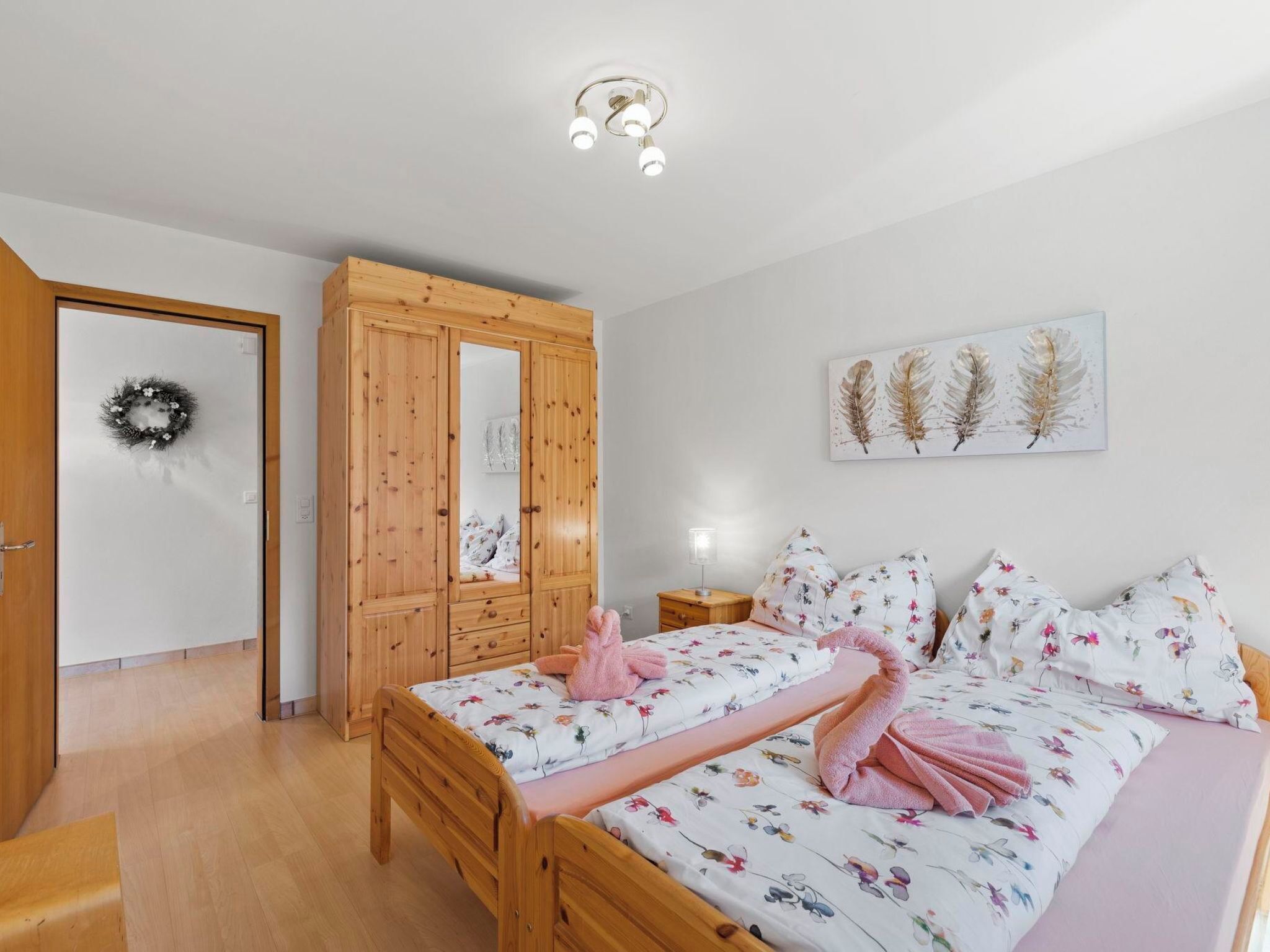 A bedroom with a wooden wardrobe, a bed with floral bedding, a wall-mounted artwork, and a ceiling light fixture.