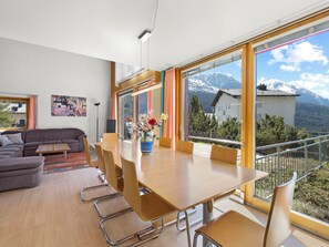 A modern living room with a sofa, a coffee table, and a dining area with a table and chairs. There is a large window with a view of mountains. - Golden Peaks Ferienhaus Ferienhaus, Dusche und Bad, WC, 4 oder mehr Schlafräume by Interhome (Valbella)