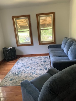 A living room with a gray sectional sofa, a patterned rug, a small black heater, and two windows with wooden frames.