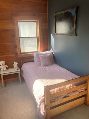A bedroom with a wooden bed, a bedside table with a teddy bear, a window, and a painting on the wall.