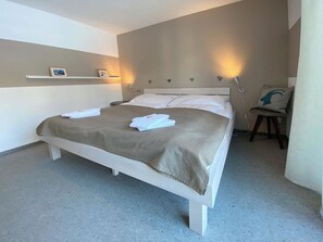 Una cama bien hecha con sábanas blancas y una manta color beige, una mesita de noche con una lámpara, y una silla con un cojín.