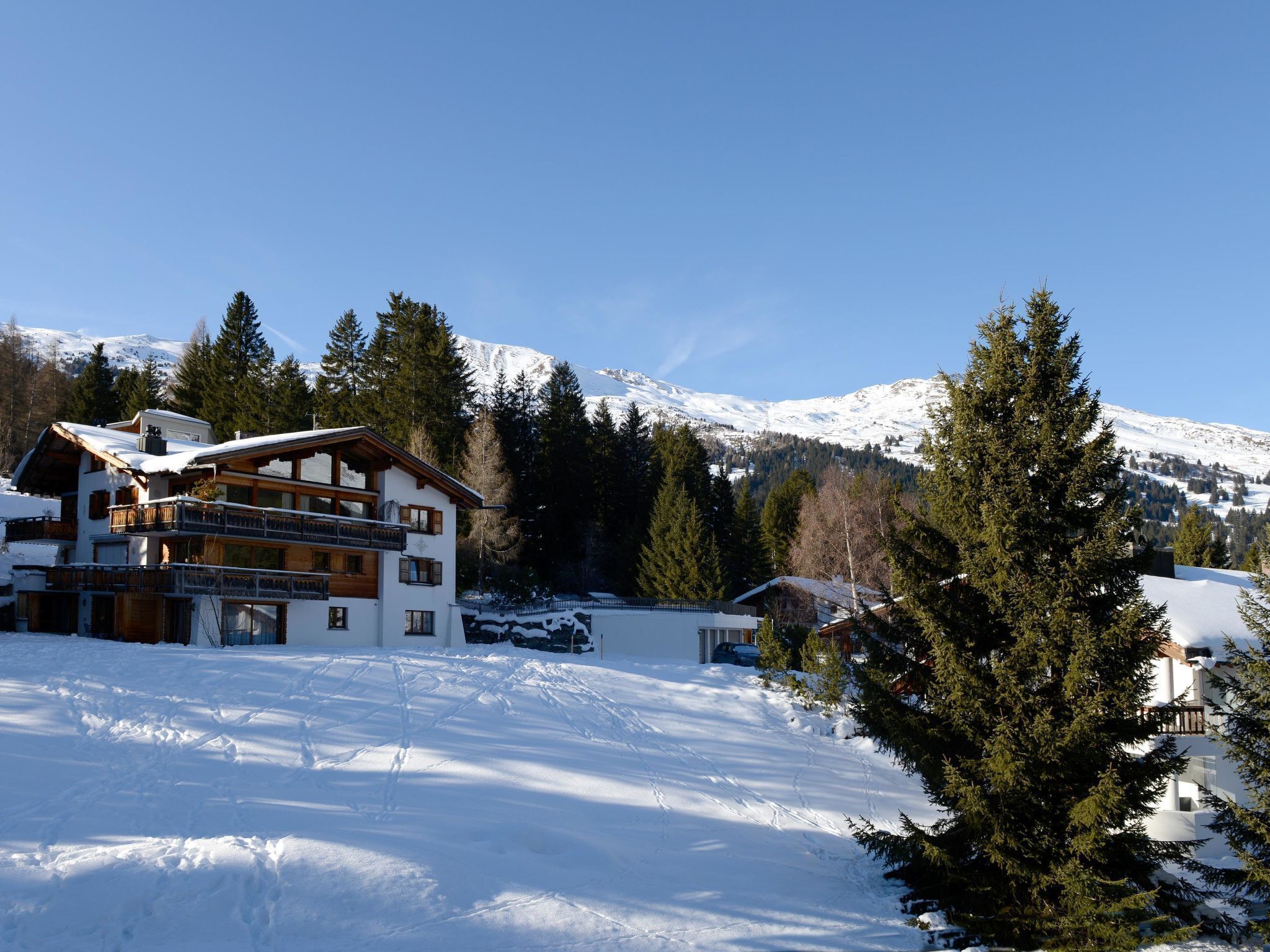 Een chalet met meerdere verdiepingen met een skilift op de achtergrond, omgeven door besneeuwde bergen en dennenbomen.