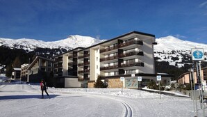 Ein mehrstöckiges Hotel mit Balkonen, eine Skipiste im Vordergrund und schneebedeckte Berge im Hintergrund.