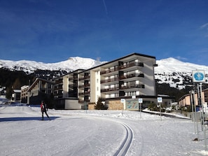 Ein mehrstöckiges Hotel mit Balkonen, eine Skipiste im Vordergrund und schneebedeckte Berge im Hintergrund.