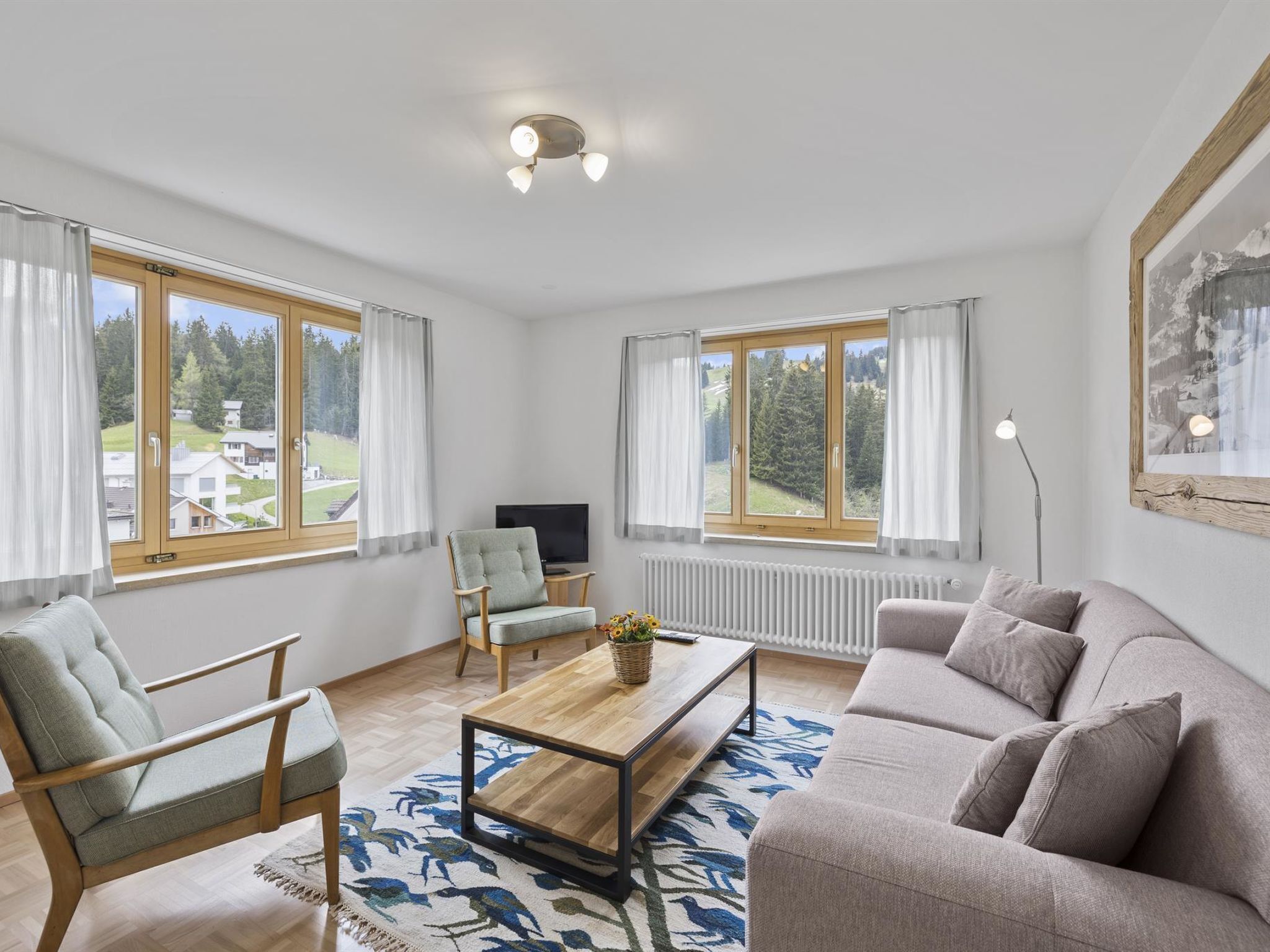 Ein modernes Wohnzimmer mit einer Couch, einem Sessel und einem Couchtisch. Große Fenster mit Vorhängen, ein Fernseher und eine Heizung sind ebenfalls vorhanden.