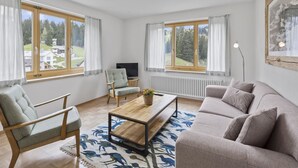 Moderni olohuone, jossa on sohva, nojatuoli ja sohvapöytä. Huoneessa on suuret ikkunat, joissa on verhot, televisio ja patteri.