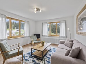 A modern living room with a sofa, armchair, and a coffee table. There are large windows with curtains, a television, and a radiator. - Bellavista 6 Appartement/Fewo, Bad, WC, 3 Schlafräume Ferienwohnung Bellavista 6 by Interhome (Parpan)