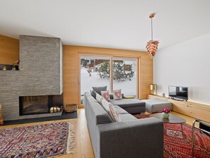 Un salon moderne avec une cheminée, un grand ensemble de canapés, un téléviseur et une vue sur un paysage enneigé, visible à travers une porte vitrée.