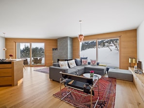 A modern living room with a large sectional sofa, a dining area with a wooden table, and a view of a snowy landscape through large windows. - Praval Appartement/Fewo Ferienwohnung Praval by Interhome (Valbella)