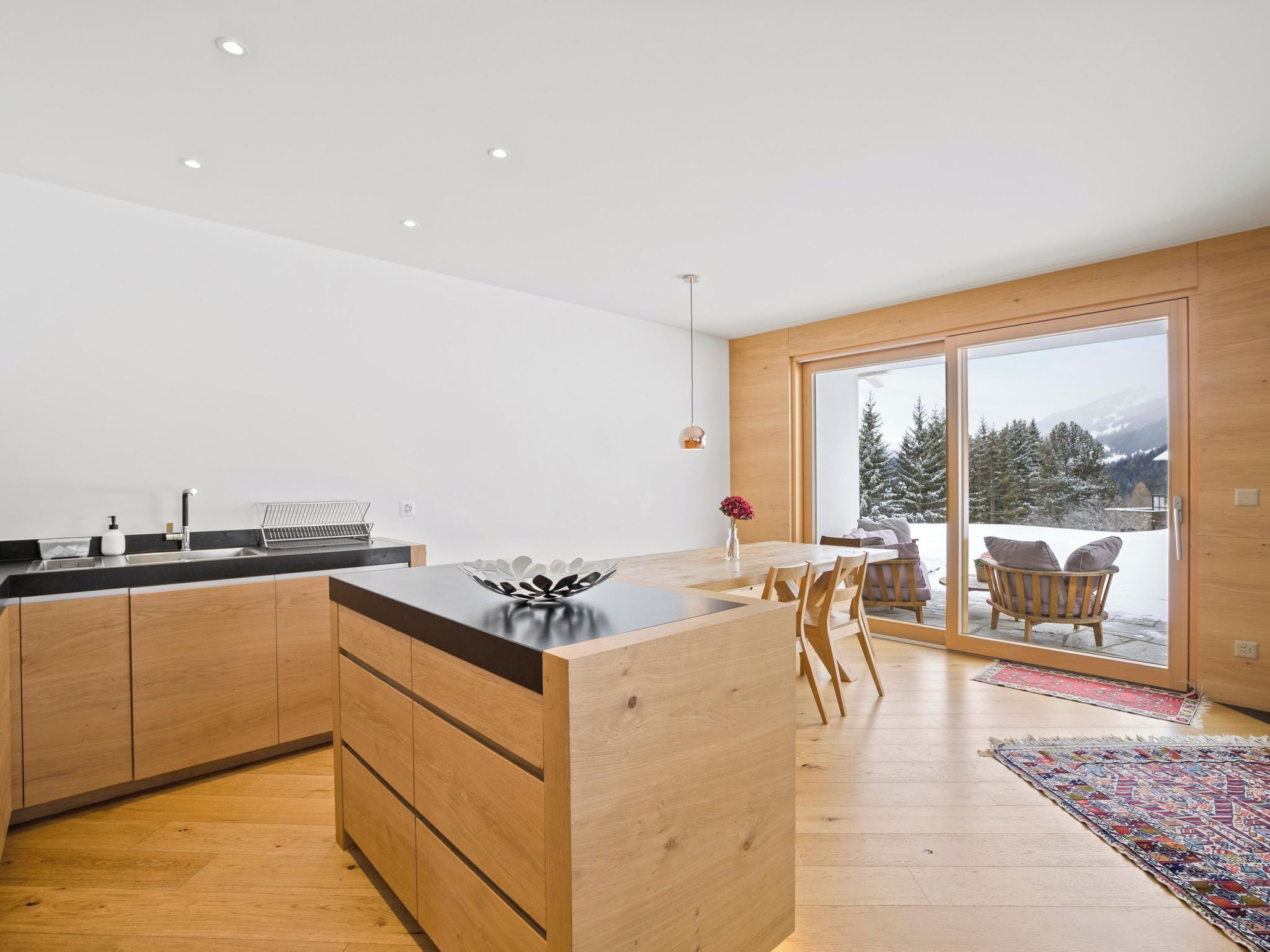 Eine moderne Küche mit Holzausstattung und einer zentralen Kochinsel, ein Essbereich mit Stühlen und eine große Schiebeglasdoor, die zu einer verschneiten Außenanlage führt.