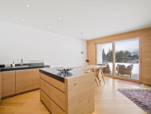 A modern kitchen with wooden cabinets and a central island, a dining area with chairs, and a large sliding glass door leading to a snowy outdoor area. - Praval Appartement/Fewo Ferienwohnung Praval by Interhome (Valbella)