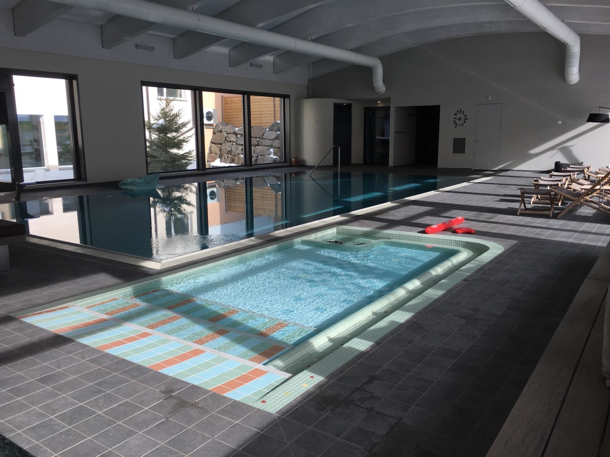 An indoor swimming pool with a tiled floor, a hot tub, and lounge chairs.