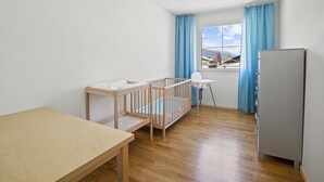 Et værelse med en rejsegrødde, en babystol, et bord og et køleskab.