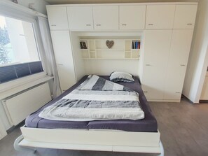 Ein Schlafzimmer mit einem Bett, einem Fenster und einem weißen Kleiderschrank mit Regalen.