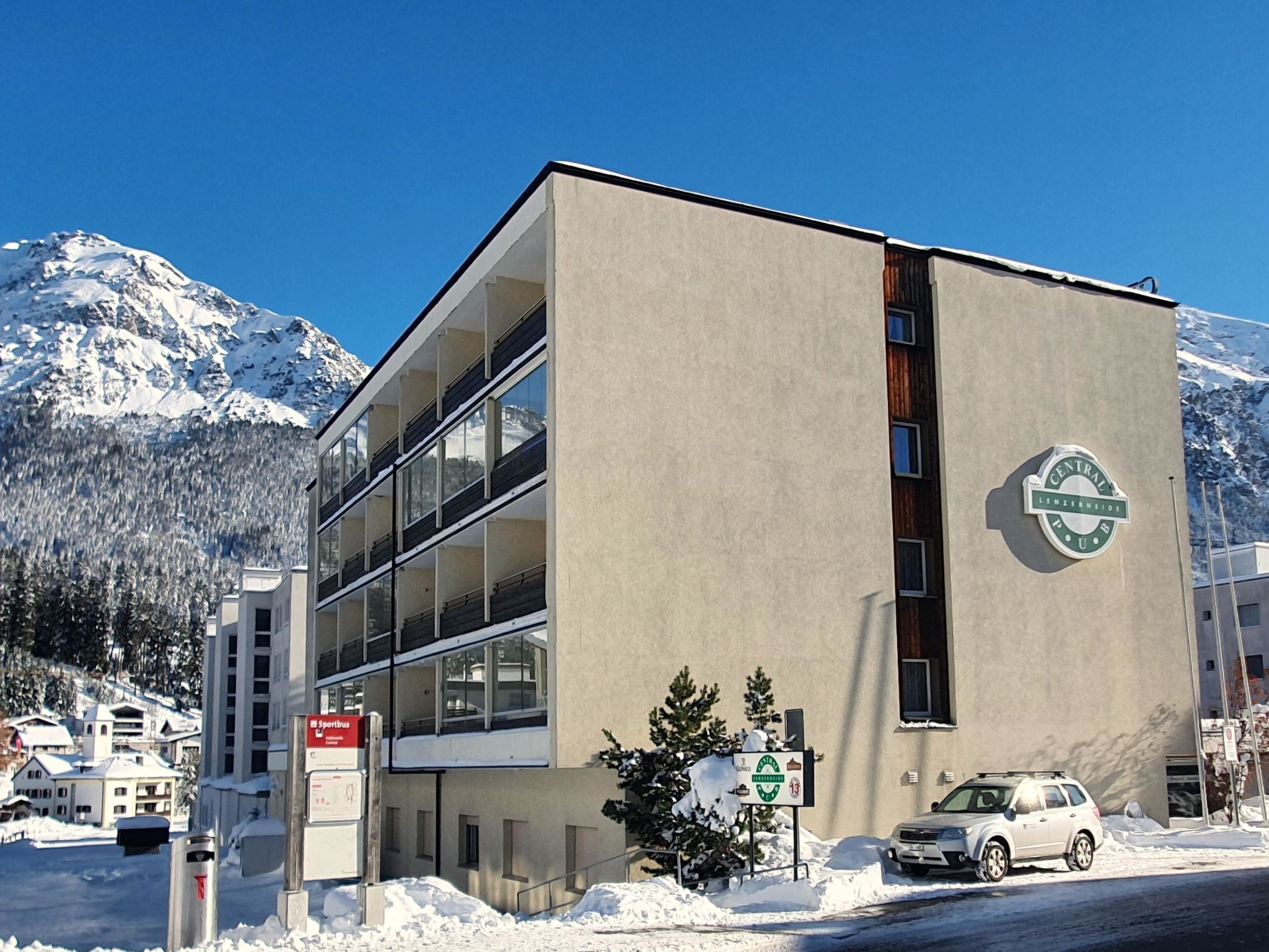 Un edificio moderno con un logotipo central, un letrero para instalaciones deportivas y un coche estacionado al frente.