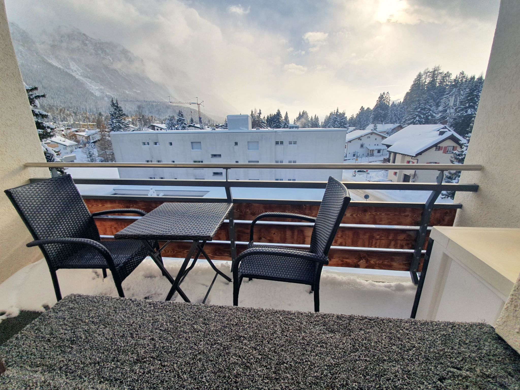 A balcony with two chairs and a table, overlooking a snowy village with buildings and mountains in the background.