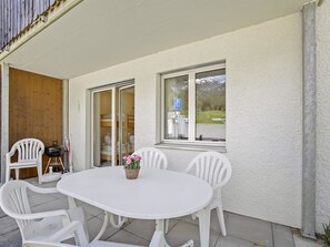 Eine Terrasse im Freien mit einem weißen Tisch und Stühlen, einem Blumenkorb und Blick auf einen Berg durch das Fenster.