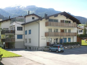 Ein mehrstöckiges Gebäude mit Balkonen, ein Auto davor und Berge im Hintergrund.