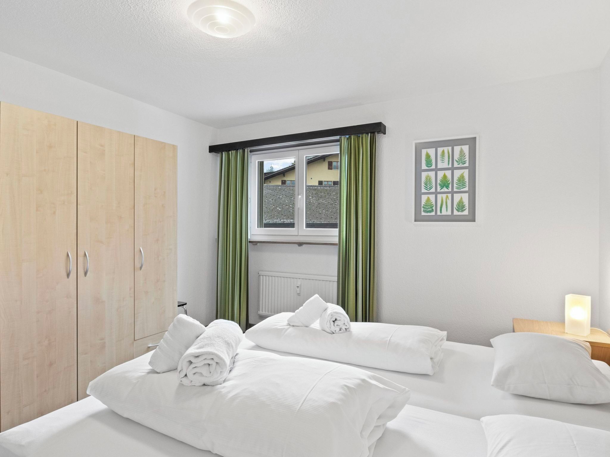 A hotel room with two beds, a wooden wardrobe, a window with curtains, and a wall-mounted picture.
