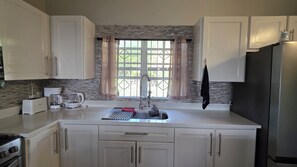 Cocina con armarios blancos, un refrigerador de acero inoxidable, una cafetera y una ventana con vista a follaje.