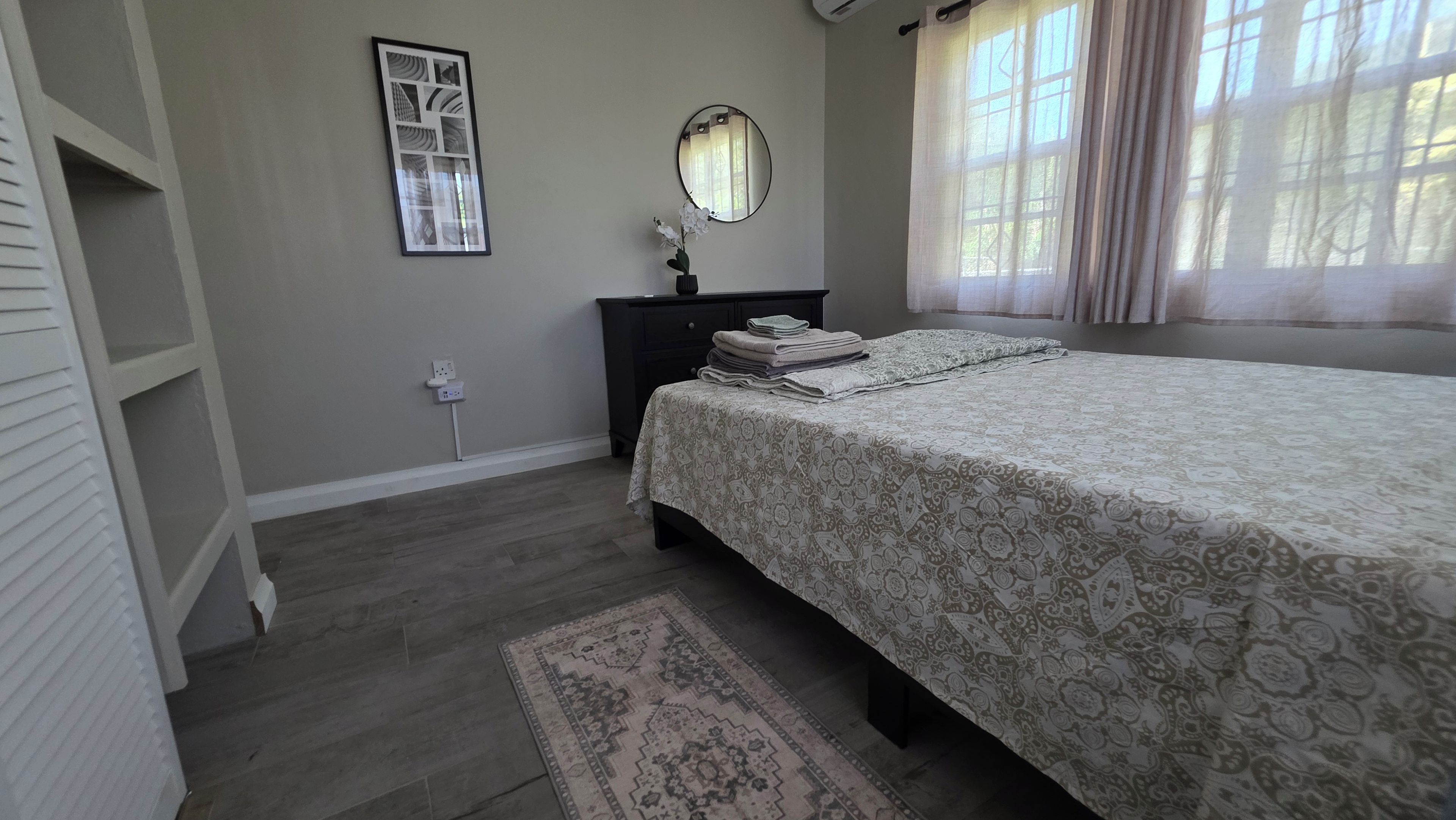Un dormitorio con cama, mesita de noche, espejo, toma de corriente y ventana con cortinas.