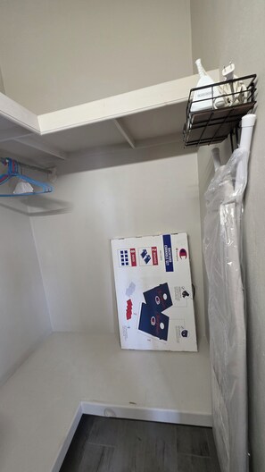 Pequeña habitación cerrada con techo blanco, una estantería metálica con diversos objetos y un objeto envuelto en plástico a la derecha.
