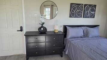 A bedroom with a dark wooden dresser, a bed with grey bedding, a round mirror, and abstract wall art.