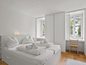 A modern bedroom with a large bed, two pillows, a wooden chair, and a window with a view of greenery. - Triangel Raber Appartement/Fewo Triangel Raber by Interhome (Parpan)