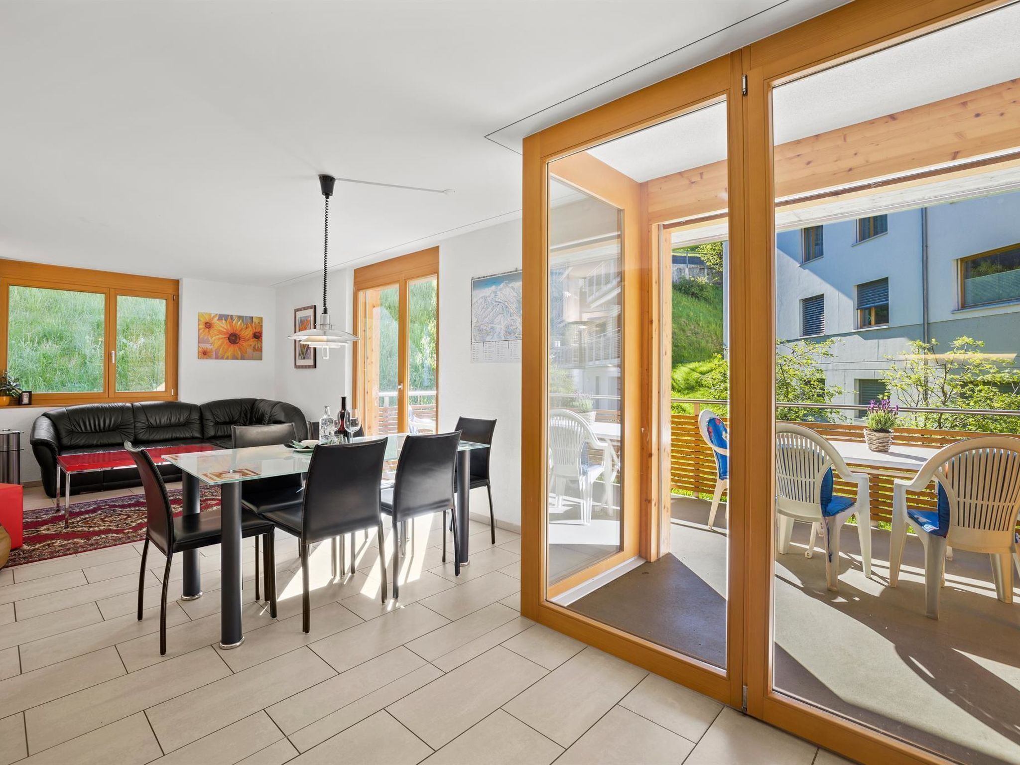 Ein modernes Wohnzimmer mit Essbereich, großen Schiebeglasfenstern, die auf eine Terrasse im Freien führen, und Blick auf ein Gebäude im Hintergrund.