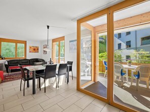 A modern living room with a dining area, large sliding glass doors leading to an outdoor patio, and a view of a building in the background. - Hemmadi Appartement/Fewo,   Ferienwohnung Hemmadi by Interhome (Churwalden)