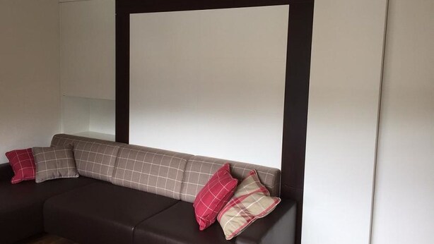A modern living room with a dark brown sofa, white walls, and a large window. - Haus Allod 201 Appartement/Fewo Allod 201 Gretler by Interhome (Lenzerheide)