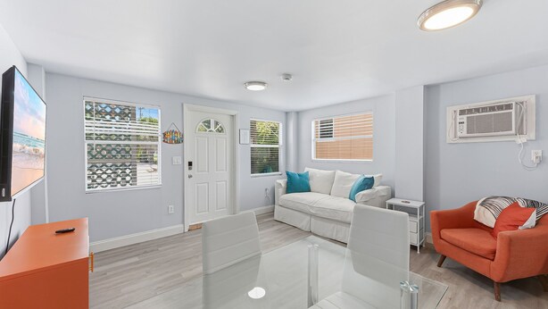 A modern living room with a glass table, white sofa, orange armchair, and a TV on a stand. - Walk To Beach, North End Restaurants, Night Life Steps Away! (Fort Myers Beach)